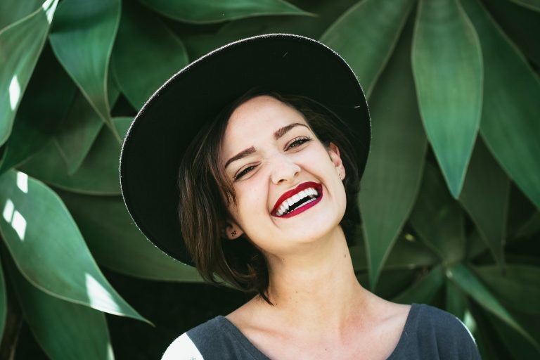 Young woman with short dark hair wearing a black hat, smiling confidently and showing bright white teeth