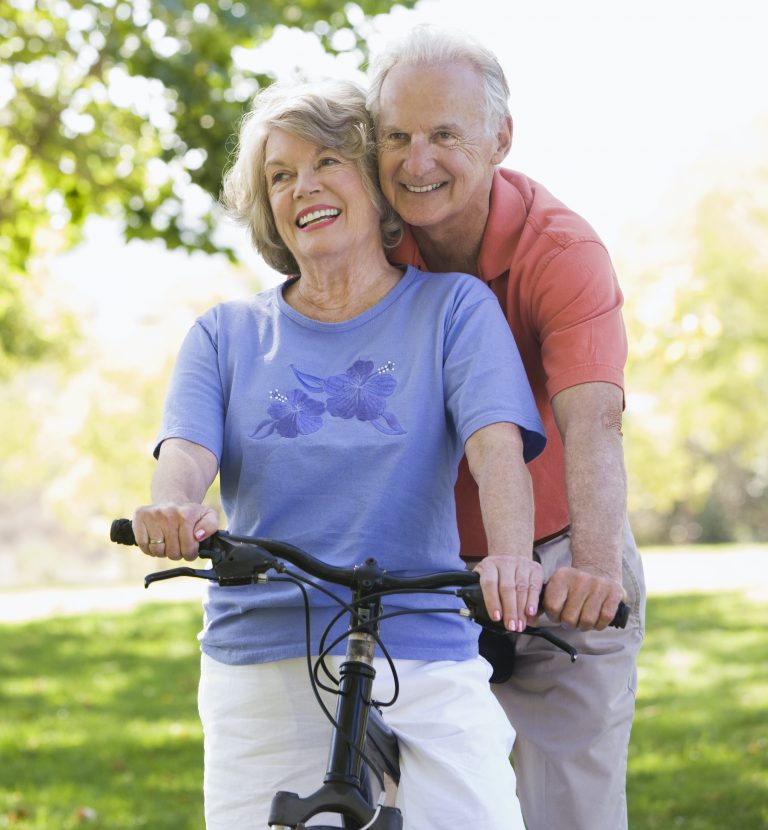 Smiling elderly couple riding a bicycle together outdoors, showing confidence with dentures
