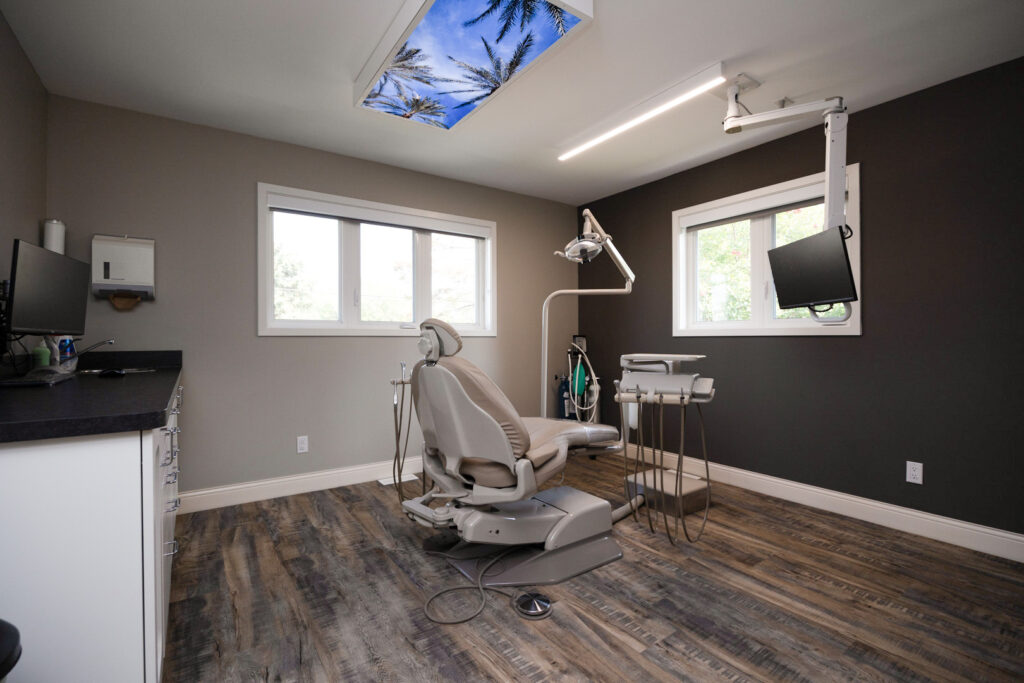 Spacious dental operatory with modern equipment, wood flooring, ceiling sky panel, and natural light from large windows