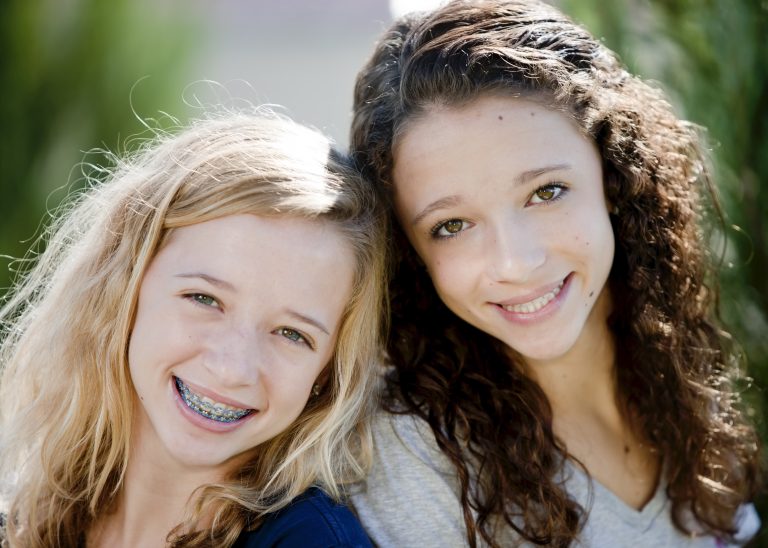 Two teenage girls smiling outdoors, one with braces and the other with straight teeth