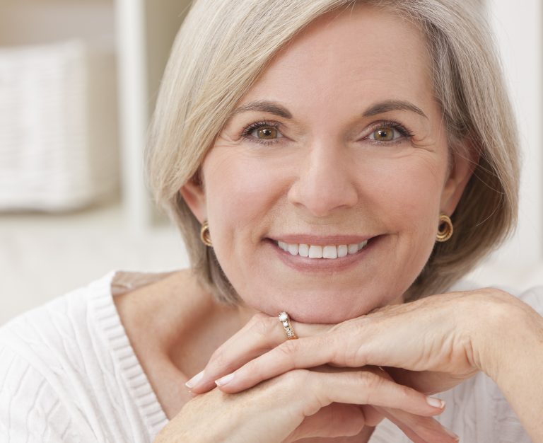 Smiling mature woman with short gray-blonde hair resting her chin on her hands, showing healthy teeth