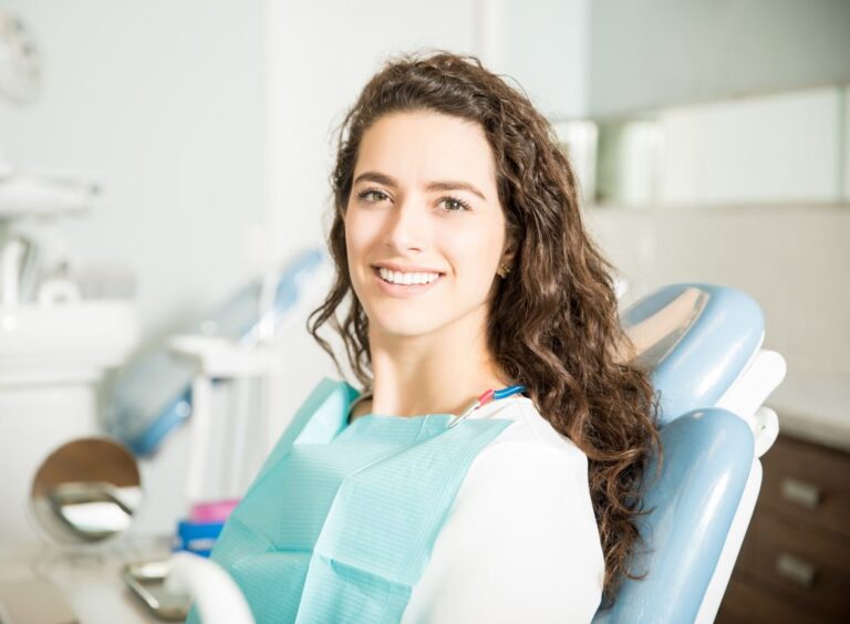 Young woman with curly brown hair smiling confidently while sitting in a dental chair wearing a patient bib