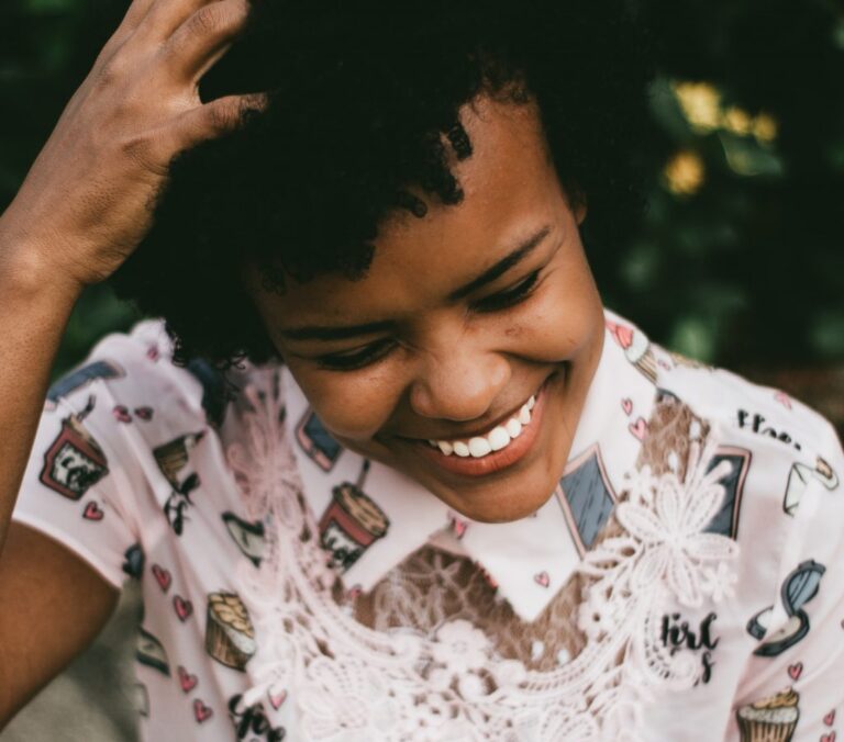 Woman smiling with bright white teeth, wearing a patterned shirt and looking down joyfully after a cosmetic dental procedure