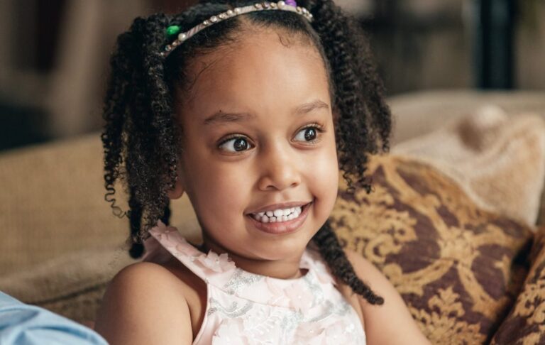 Smiling young girl with curly hair and beaded headband sitting on a couch, showing clean white teeth