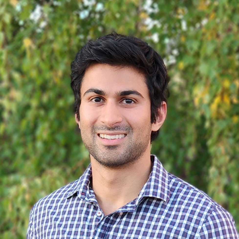 Dr. Rehan Merchant smiling outdoors in a checkered shirt with greenery in the background
