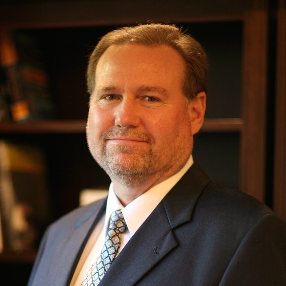 Portrait of Dr. Everett K. Postill in a suit, standing in front of a bookshelf at Orchard Family Dental